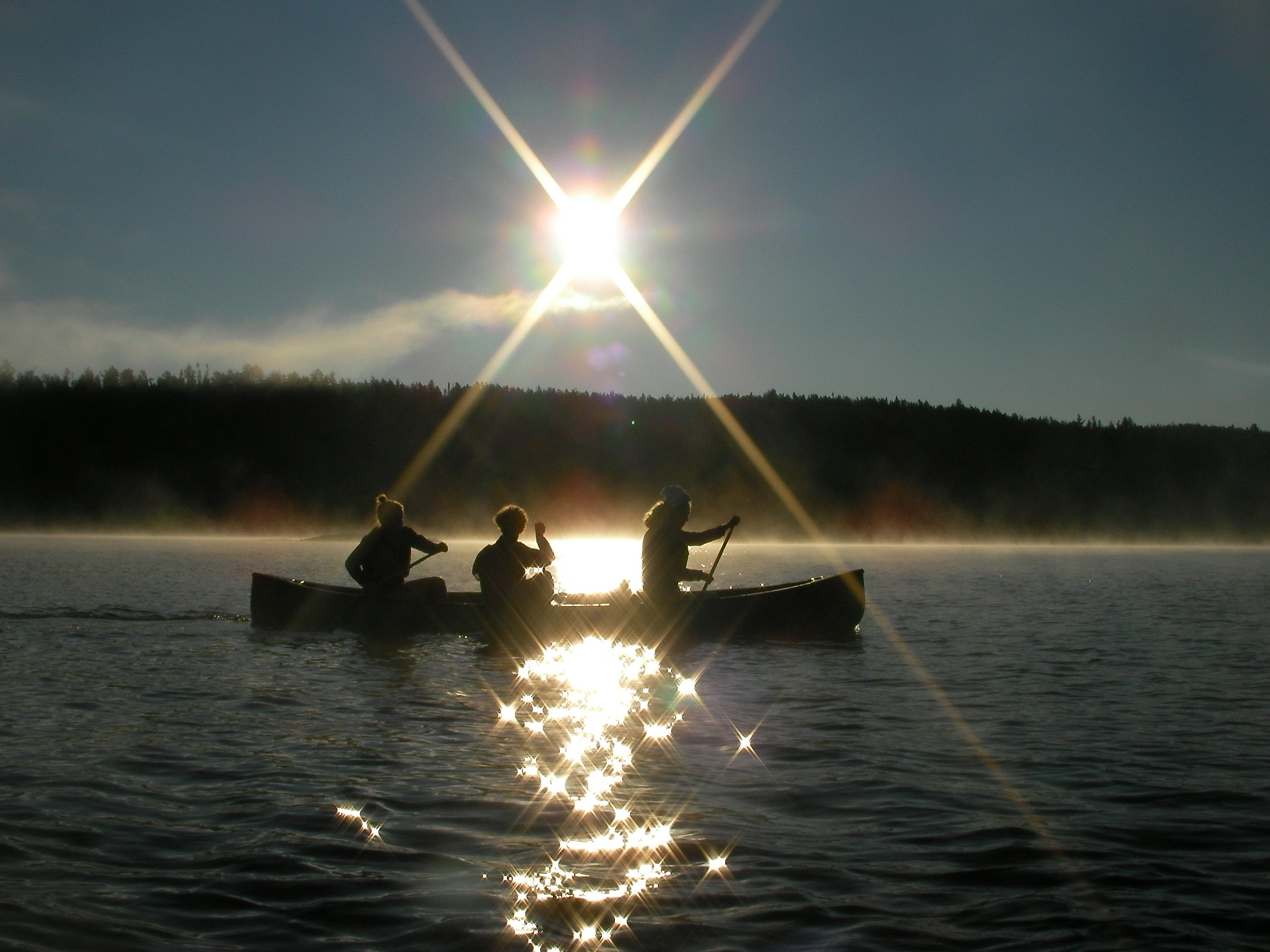 A group of three students canoeing on a lake with the sun in the background